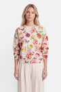 Eine Frau mit blonden Haaren trägt einen cremefarbenen Sweater mit einem durchgehenden Print aus bunten Blumen und Schmetterlingen. Sie steht mit neutralem Gesichtsausdruck nach vorne gerichtet da. Außerdem trägt sie eine hellbeige Hose, ein goldenes Armband und schlichte Ohrringe. Der Hintergrund ist schlicht weiß.
