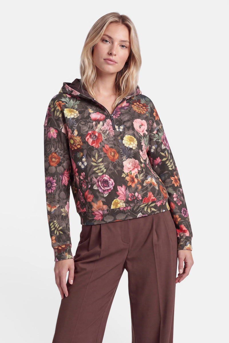 Eine Person steht da und trägt einen braunen Sweat-Hoodie mit Blumenmuster, der mit einem durchgehenden Allover-Print aus leuchtenden Blumen in Rosa, Rot und Gelb verziert ist. Der aus weicher Baumwolle gefertigte Hoodie mit Reißverschluss verfügt über eine Kapuze und wird mit einer passenden braunen Hose kombiniert. Die Person hat blondes Haar und einen neutralen Gesichtsausdruck und steht vor einem schlichten weißen Hintergrund.
