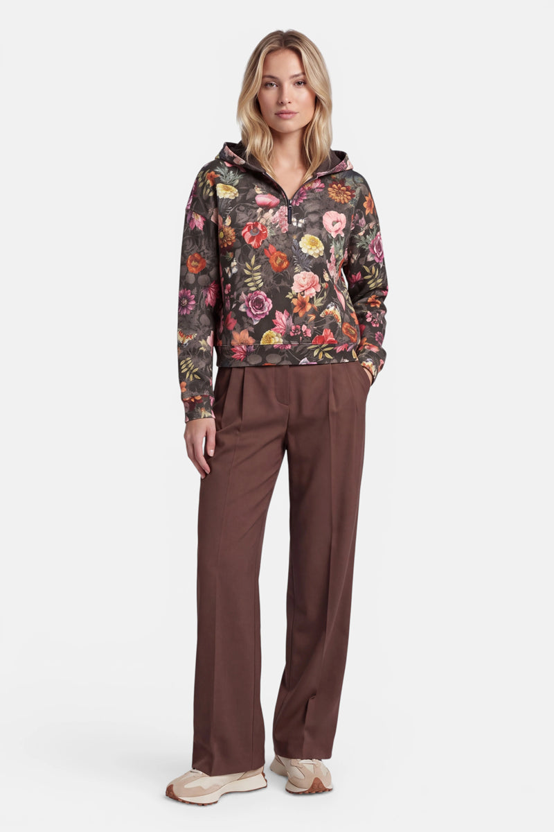 Eine Frau steht mit dem Blick nach vorne und trägt einen dunklen Sweat-Hoodie mit durchgehendem Blumenmuster und Reißverschluss aus Baumwolle, dazu eine braune Hose mit weitem Bein und beigefarbene Sneaker. Ihre Hände sind entspannt, eine steckt in der Tasche. Sie hat schulterlanges blondes Haar und einen neutralen Gesichtsausdruck. Der Hintergrund ist schlicht weiß.