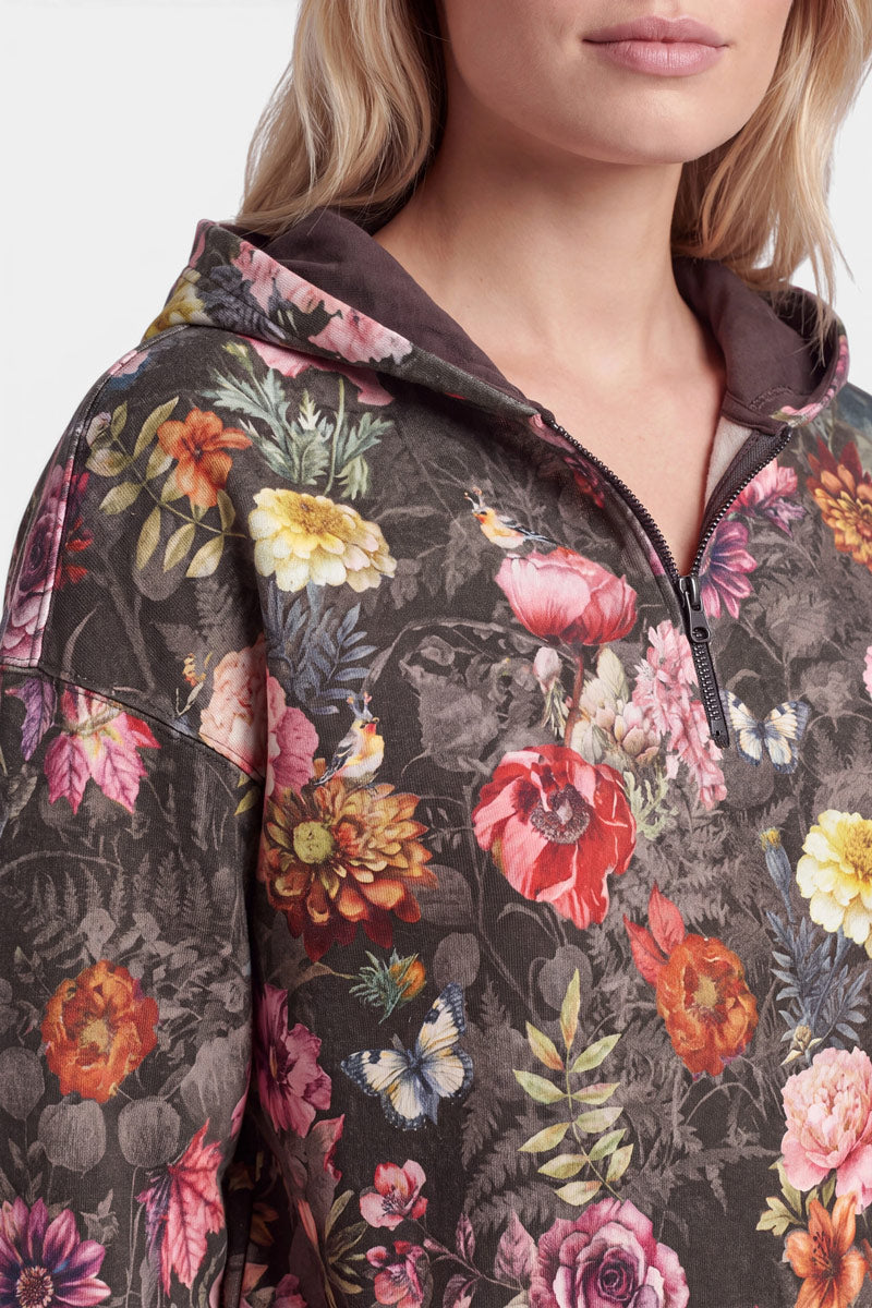 Eine Person, die einen stilvollen Sweat-Hoodie aus weicher Baumwolle trägt, verziert mit einem lebhaften Allover-Print aus Rosen, Pfingstrosen und anderen Blumen in Rot-, Gelb- und Rosatönen, ergänzt durch grüne Blätter und einen zarten Schmetterling – alles vor einem dunklen Hintergrund. Nur der untere Teil des Gesichts und der obere Oberkörper sind zu sehen.