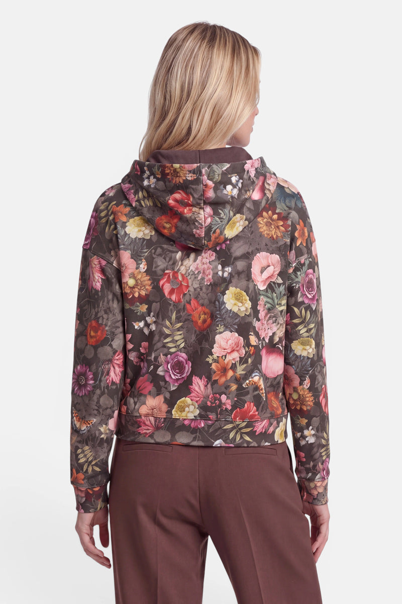Eine Frau, die mit dem Rücken zur Kamera steht, trägt einen braunen Sweat-Hoodie mit Blumenmuster aus weicher Baumwolle, der mit einem durchgehenden Print aus großen, bunten Blumen verziert ist, dazu eine braune Hose. Ihr blondes Haar trägt sie offen und sie steht vor einem schlichten weißen Hintergrund. Der Hoodie hat eine lockere Passform und eine sichtbare Kapuze.