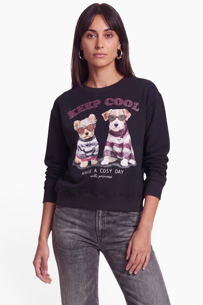 Eine Frau mit glattem braunem Haar trägt ein schwarzes Sweatshirt mit Hundemotiv, auf dem zwei illustrierte Hunde in Pullovern abgebildet sind, über denen der Schriftzug „KEEP COOL“ und darunter „HAVE A COSY DAY“ mit einer Prinzessin zu sehen ist. Das Sweatshirt ist aus weicher Baumwolle gefertigt und bietet höchsten Tragekomfort. Sein verspieltes Design wird durch dezente Strass-Details unterstrichen. Sie steht vor einem schlichten weißen Hintergrund und trägt eine graue Jeans.