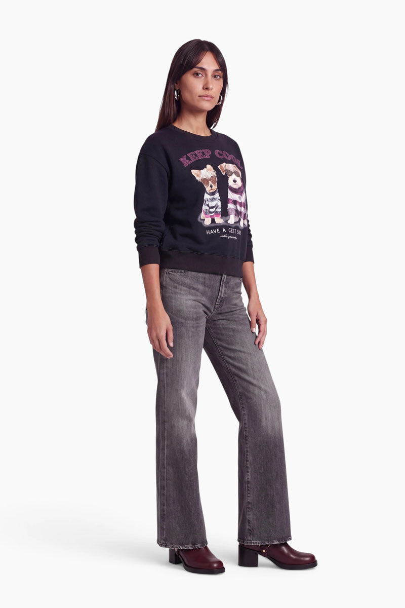 Eine Frau steht vor einem weißen Hintergrund und trägt ein schwarzes Baumwoll-Sweatshirt mit einem Hundemotiv und Schriftzug, eine graue Jeans mit weitem Bein sowie dunkle Stiefel mit Absatz. Sie hat glattes, dunkles Haar und einen neutralen Gesichtsausdruck; ihre Arme liegen entspannt an den Seiten.