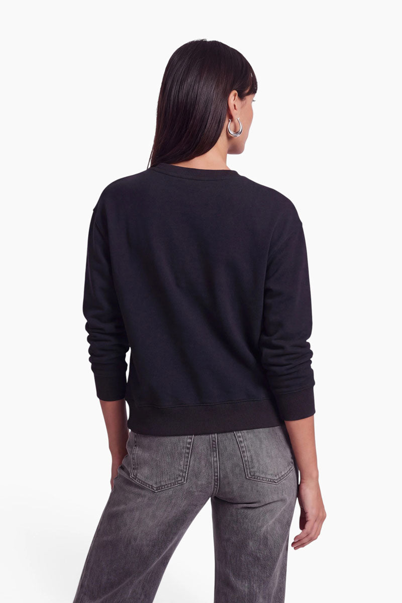 Eine Frau mit glattem, dunklem Haar, die große Creolen, ein schwarzes, langärmeliges Baumwoll-Sweatshirt und eine graue Jeans mit hoher Taille trägt, steht mit dem Rücken zur Kamera vor einem schlichten weißen Hintergrund.