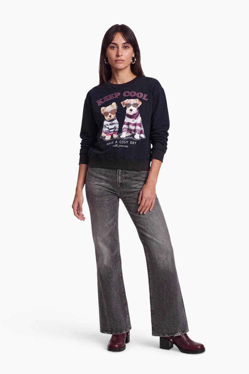 Eine Frau steht da und trägt ein schwarzes „Hundeprint“-Sweatshirt aus Baumwolle mit aufgedruckten Hunden und dezenten Strass-Akzenten, dazu eine graue Schlagjeans und burgunderrote Stiefel mit Absatz. Sie hat glattes dunkles Haar, Creolen und einen ausdruckslosen Gesichtsausdruck und steht vor einem schlichten weißen Hintergrund.