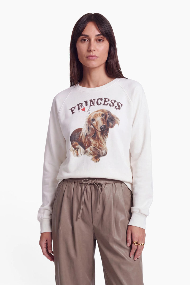 Eine Frau mit glattem braunem Haar trägt ein weißes Baumwoll-Sweatshirt mit einem niedlichen Dackel-Aufdruck und der Aufschrift „PRINCESS“ darüber. Außerdem trägt sie eine taupefarbene Hose mit Kordelzug und mehrere Ringe. Sie steht vor einem schlichten weißen Hintergrund und blickt direkt in die Kamera.