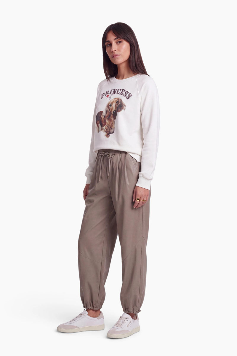 Eine Frau steht vor einem weißen Hintergrund und trägt ein weißes Baumwoll-Sweatshirt mit der Aufschrift „PRINCESS“ und einem niedlichen Dackel-Aufdruck, eine taupefarbene Jogginghose sowie weiße Sneaker. Sie hat langes, glattes, dunkles Haar und einen neutralen Gesichtsausdruck; ihre Hände stecken in den Taschen.