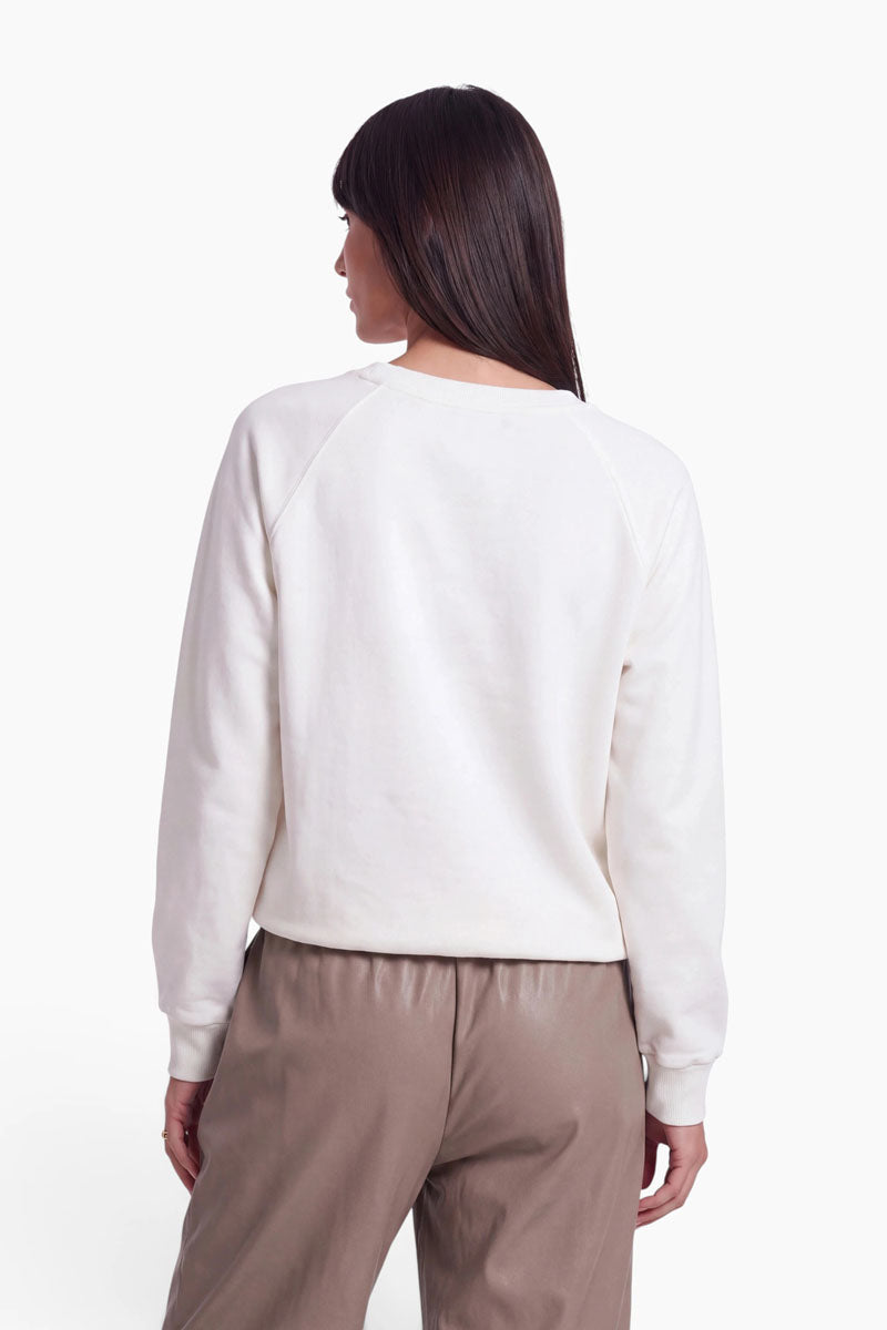 Eine Frau mit glattem braunem Haar steht mit dem Rücken zur Kamera. Sie trägt ein cremefarbenes Baumwoll-Sweatshirt mit einem dezenten Dackel-Aufdruck und langen Ärmeln, dazu eine taupefarbene Hose mit hoher Taille. Der Hintergrund ist schlicht weiß.