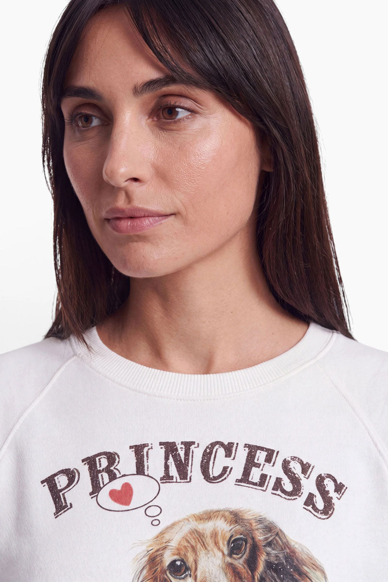 Eine Frau mit glattem braunem Haar und Pony blickt leicht nach links. Sie trägt ein weißes Baumwoll-Sweatshirt mit einem niedlichen Dackel-Aufdruck und dem Wort „PRINCESS“ in großen Vintage-Buchstaben, über dem Hund befindet sich ein rotes Herz in einer Sprechblase. Der Hintergrund ist weiß.