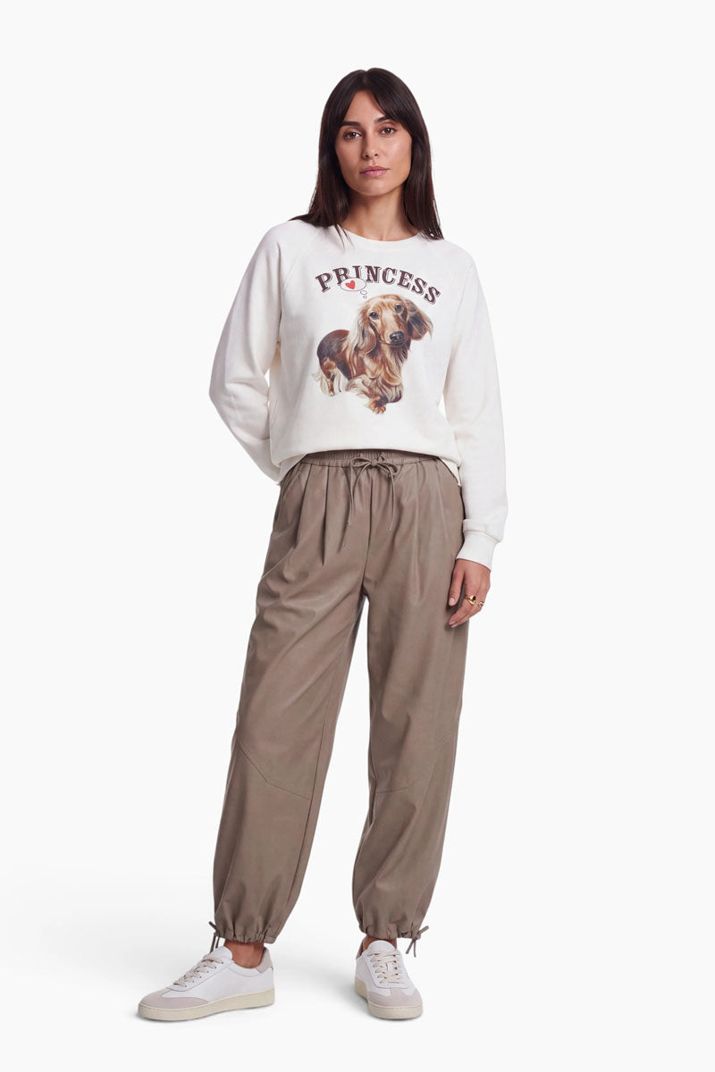 Eine Frau steht da und trägt ein weißes Baumwoll-Sweatshirt mit der Aufschrift „PRINCESS“ und einem niedlichen Dackel-Aufdruck, eine lockere, taupefarbene Jogginghose sowie weiße Sneaker. Sie hat glattes, dunkles Haar und einen neutralen Gesichtsausdruck und steht vor einem schlichten weißen Hintergrund.