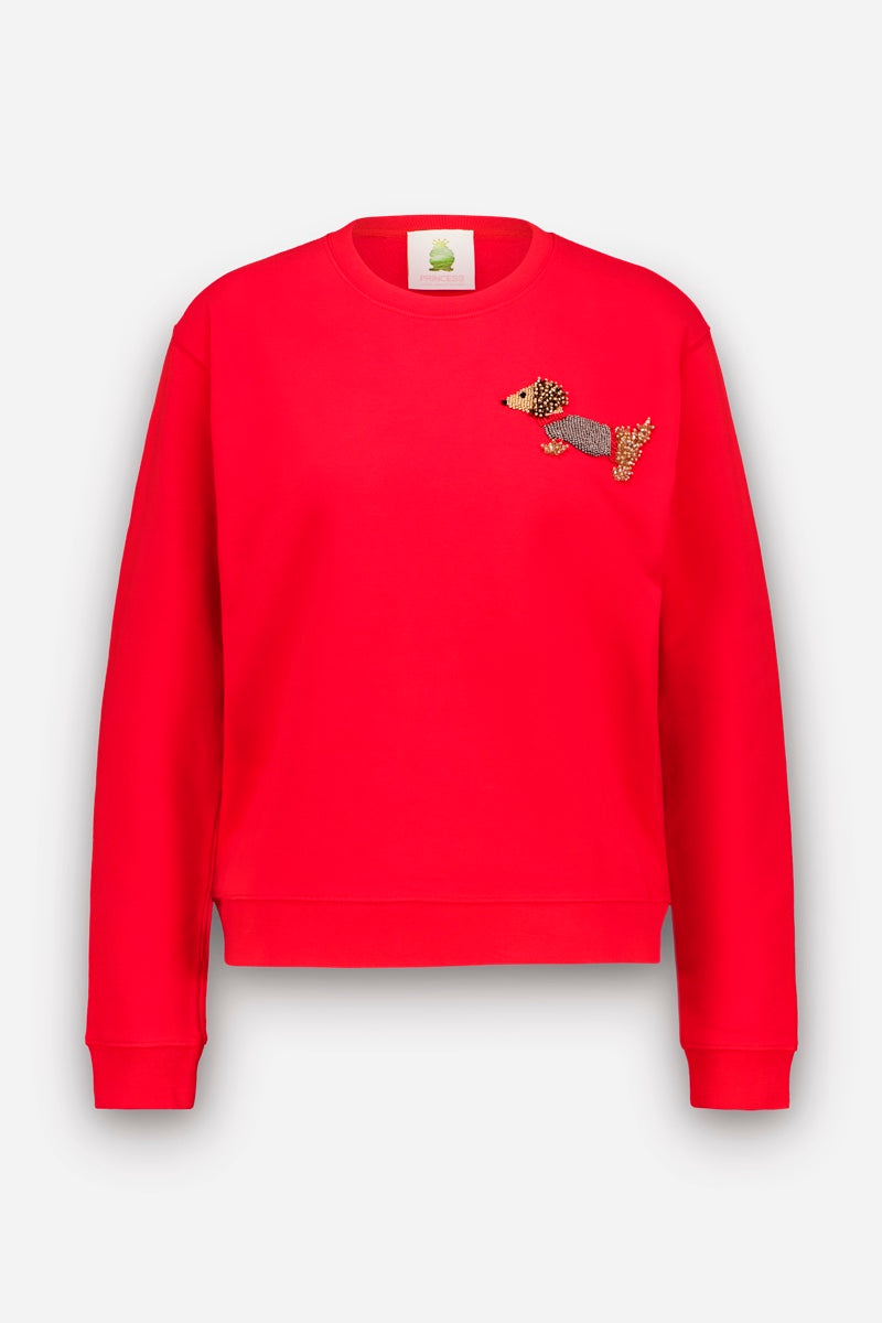 Leuchtend rotes Sweatshirt mit langen Ärmeln, Rundhalsausschnitt und geripptem Saum. Auf der linken Brust befindet sich ein gestickter Dackel-Applikationsaufnäher, bei dem für den Körper, das Ohr und den Schwanz des Hundes verschiedene Farben und Muster verwendet wurden. Das Sweatshirt aus Baumwolle ist vor einem schlichten weißen Hintergrund abgebildet.