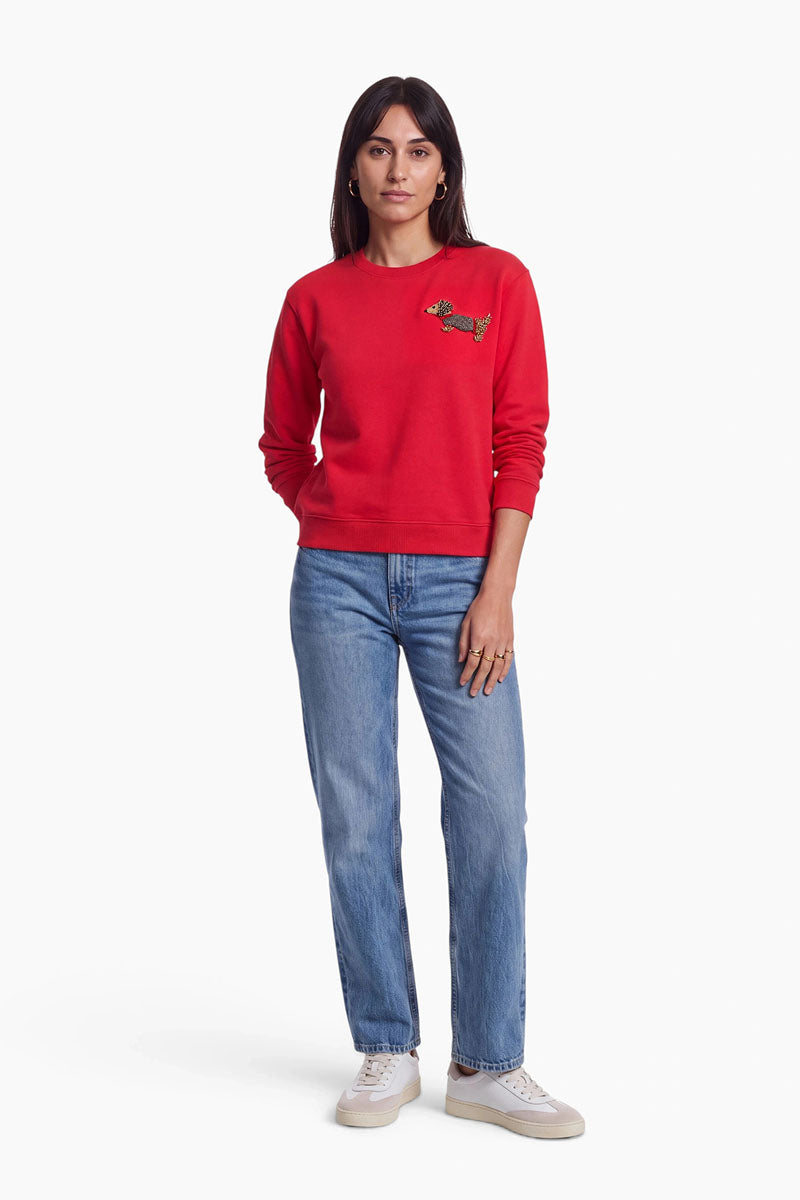Eine Frau steht mit dem Gesicht nach vorne und trägt ein rotes Sweatshirt aus Baumwolle mit Rundhalsausschnitt und einer kleinen Dackel-Applikation auf der Brust, dazu hellblaue Jeans und weiße Sneaker. Sie hat glattes dunkles Haar, trägt Creolen und hat einen neutralen Gesichtsausdruck. Der Hintergrund ist schlicht weiß.