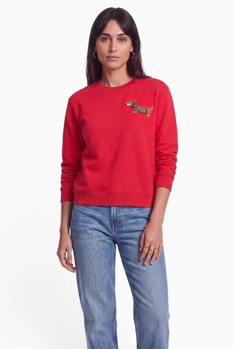Eine Frau mit langen braunen Haaren trägt ein rotes Sweatshirt aus Baumwolle mit einer kleinen gestickten Dackelapplikation auf der linken Brustseite sowie hellblaue Jeans. Das Sweatshirt hat einen klassischen Rundhalsausschnitt. Sie steht vor einem schlichten weißen Hintergrund und blickt mit neutralem Gesichtsausdruck direkt in die Kamera.