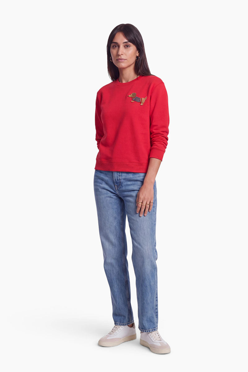 Eine Frau steht mit dem Blick nach vorne und trägt ein rotes Baumwolle-Sweatshirt mit einem kleinen Fuchs-Aufnäher auf der linken Brust, hellblaue Jeans mit geradem Bein und weiße Sneaker. Sie hat glattes, dunkles Haar und einen neutralen Gesichtsausdruck. Der Hintergrund ist schlicht weiß.