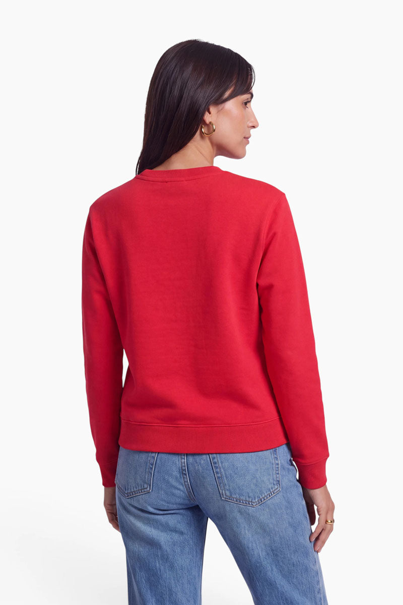 Eine Frau steht mit dem Rücken zur Kamera und trägt ein leuchtend rotes, langärmeliges Baumwoll-Sweatshirt sowie hellblaue Jeans. Ihr dunkles, glattes Haar trägt sie offen, und sie hat goldene Creolen. Der Hintergrund ist schlicht weiß, wodurch die leuchtende Farbe und die verspielte Dackelapplikation auf ihrer Kleidung besonders gut zur Geltung kommen.