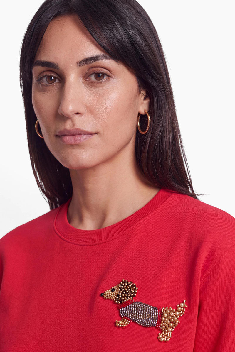 Eine Frau mit glattem, dunklem Haar und goldenen Creolen trägt ein rotes Baumwolle-Sweatshirt, das auf der Brust mit einer Brosche in Form eines Dackels mit Perlenapplikation verziert ist. Mit ausdruckslosem Gesicht blickt sie geradeaus. Der Hintergrund ist schlicht weiß.