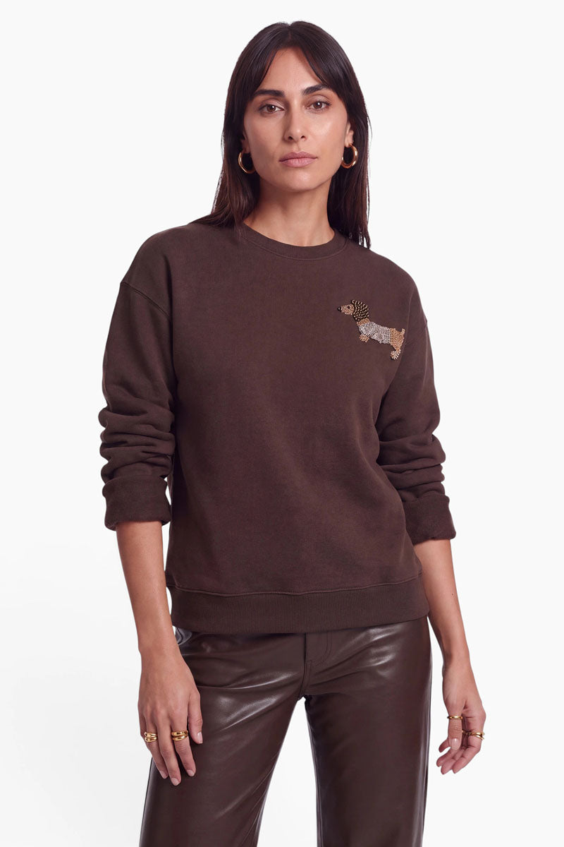 Eine Frau mit langen, dunklen Haaren trägt ein dunkelbraunes Sweatshirt aus Baumwolle mit Rundhalsausschnitt und einer kleinen Dackel-Applikation auf der linken Brust, dazu braune Lederhosen. Sie steht frontal vor einem weißen Hintergrund, die Arme locker an den Seiten hängend, und blickt direkt in die Kamera.