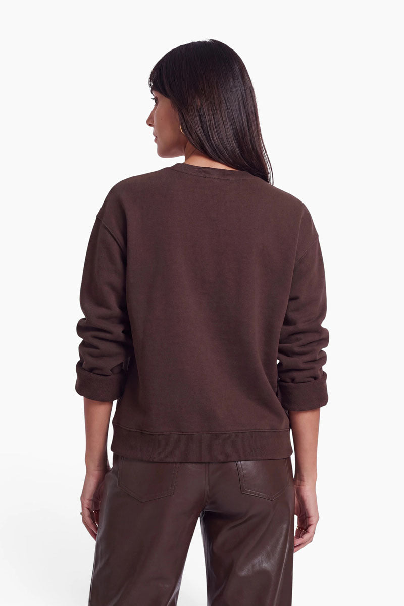 Eine Frau mit glattem, dunklem Haar steht mit dem Rücken zur Kamera. Sie trägt ein locker sitzendes braunes Sweatshirt aus Baumwolle mit Rundhalsausschnitt, dessen Ärmel lässig hochgekrempelt sind, sowie eine dazu passende braune Lederhose. Der Hintergrund ist schlicht weiß, wodurch die Farbe und die Textur des Outfits besonders zur Geltung kommen.