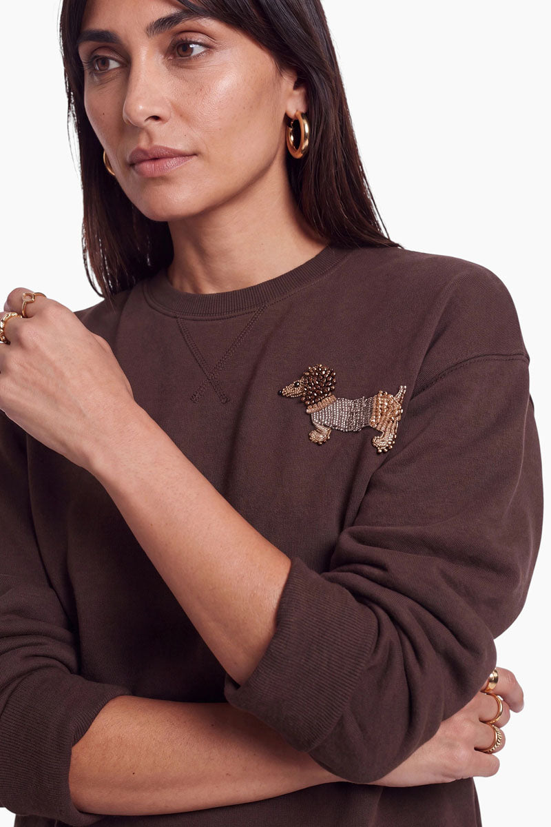Eine Frau mit langen dunklen Haaren, die ein braunes Sweatshirt aus Baumwolle mit Rundhalsausschnitt und einer perlenbesetzten Dackel-Applikation auf der Brust sowie goldene Creolen und mehrere goldene Ringe trägt, steht vor einem weißen Hintergrund und blickt mit ausdruckslosem Gesichtsausdruck zur Seite.