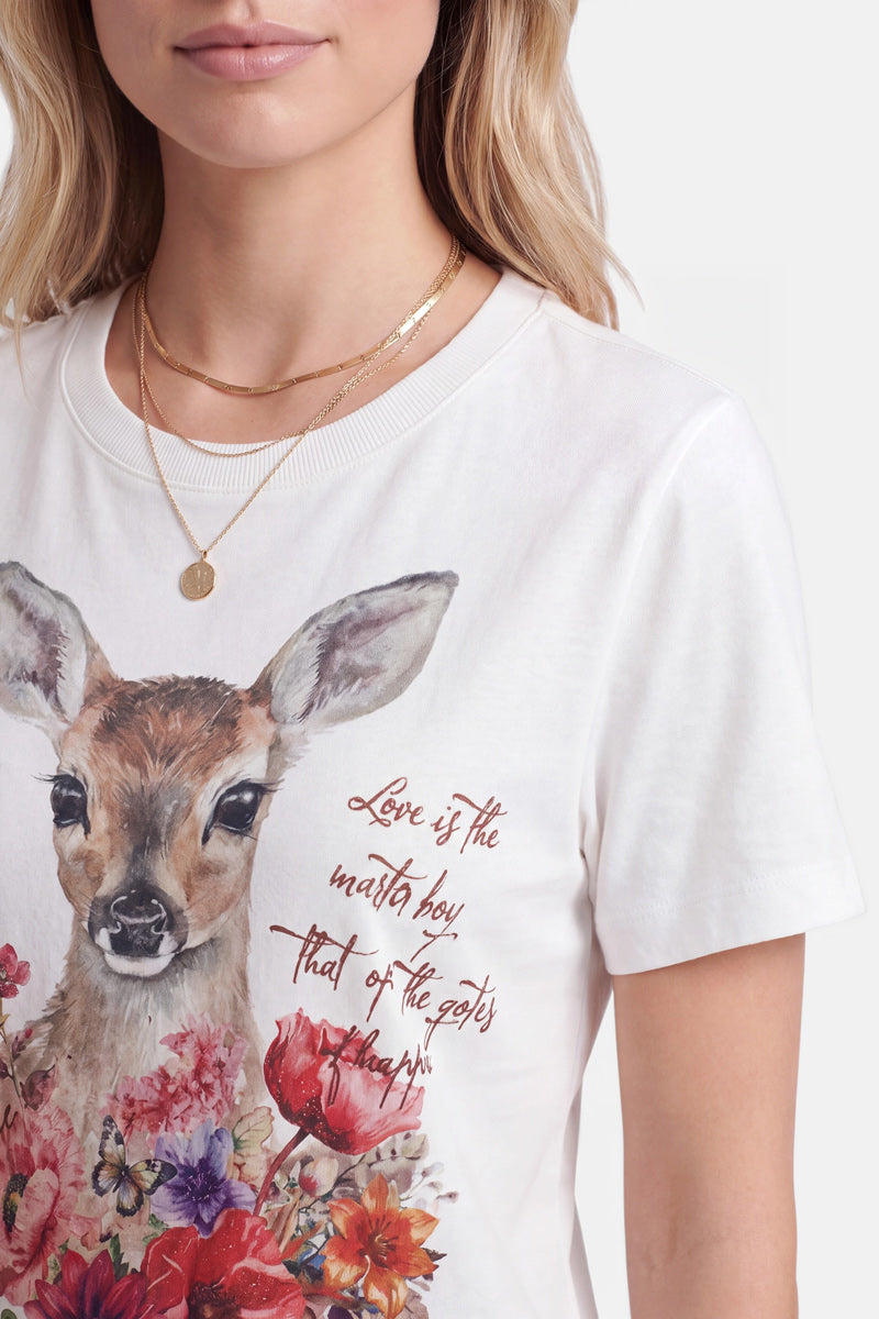 Eine Frau trägt ein weißes Baumwoll-T-Shirt mit einem detailreichen Reh-Aufdruck und bunten Blumen. Das Shirt hat einen Rundhalsausschnitt und einen in Schreibschrift gehaltenen Schriftzug, der lautet: „Liebe ist der Generalschlüssel, der die Tore zum Glück öffnet.“ Dazu trägt sie mehrere goldene Halsketten übereinander.