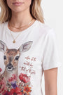 Eine Frau trägt ein weißes Baumwoll-T-Shirt mit einem detailreichen Reh-Aufdruck und bunten Blumen. Das Shirt hat einen Rundhalsausschnitt und einen in Schreibschrift gehaltenen Schriftzug, der lautet: „Liebe ist der Generalschlüssel, der die Tore zum Glück öffnet.“ Dazu trägt sie mehrere goldene Halsketten übereinander.