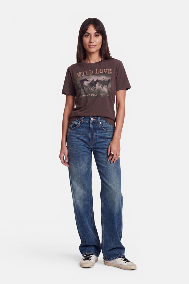 Eine Frau steht vor einem weißen Hintergrund und trägt ein braunes „Wild Love“-T-Shirt mit dezenter Strassverzierung, eine lockere blaue Jeans und weiße Sneaker. Sie hat glattes braunes Haar, die Arme locker an den Seiten und einen neutralen Gesichtsausdruck; ihr Blick ist nach vorne gerichtet.