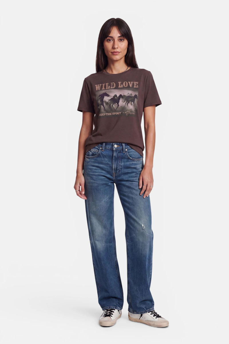 Eine Frau steht vor einem weißen Hintergrund und trägt ein braunes „Wild Love“-T-Shirt mit Pferde-Print und dezenter Strassverzierung, dazu eine locker sitzende blaue Jeans mit leichten Verwaschungen und klassische weiße Sneaker. Sie hat glattes dunkles Haar und einen neutralen Gesichtsausdruck, die Arme hält sie an den Seiten.