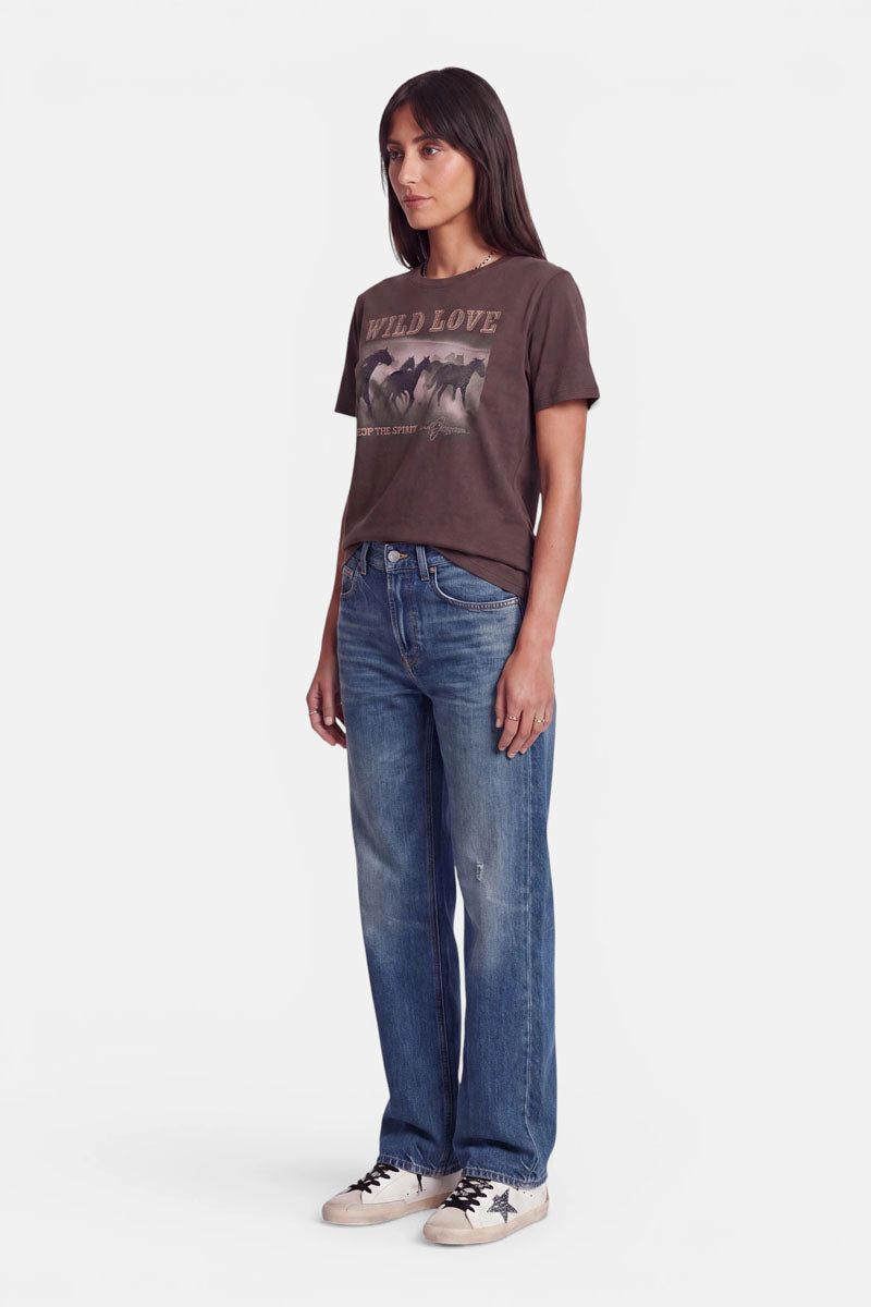Eine Frau steht mit dem Blick nach links und trägt ein braunes „Wild Love“-Shirt mit einem markanten Pferde-Print, eine blaue Jeans mit geradem Bein sowie weiße Sneaker mit schwarzen Akzenten. Sie hat glattes, dunkles Haar und einen neutralen Gesichtsausdruck. Das Baumwoll-T-Shirt verleiht ihrem lässigen Look vor dem schlichten weißen Hintergrund eine bequeme Note.