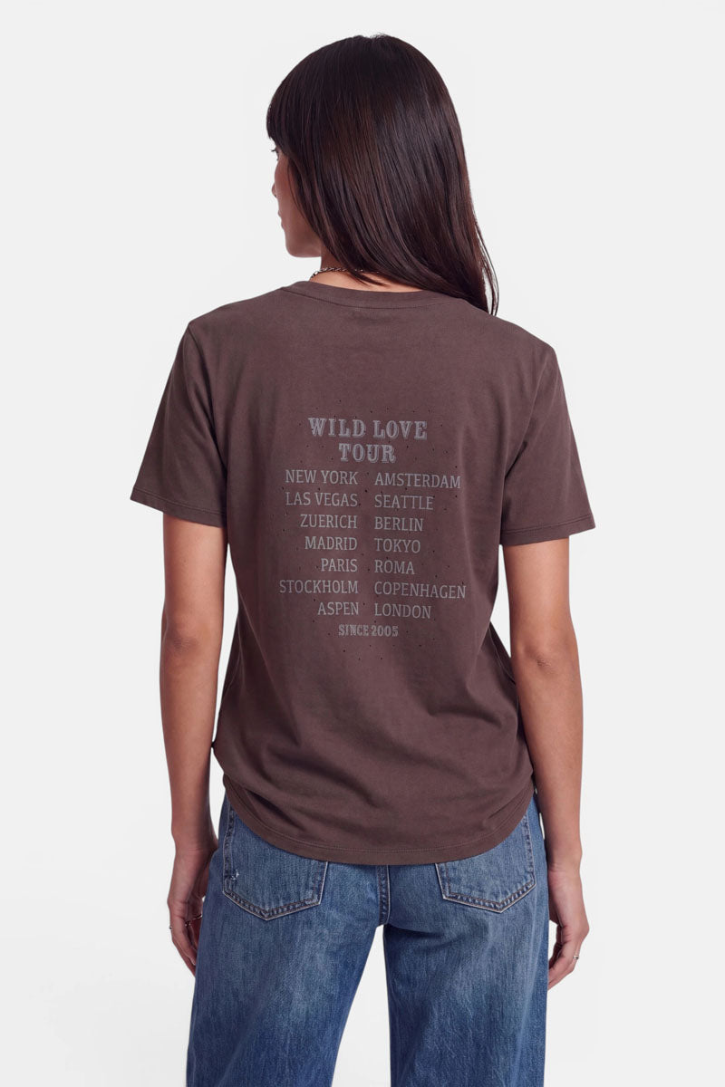 Eine Frau mit langen, dunklen Haaren trägt ein braunes „Wild Love“-T-Shirt und blaue Jeans. Auf der Rückseite des Shirts ist „Wild Love Tour“ aufgedruckt, zusammen mit einer Liste wichtiger Städte in hellgrauer Schrift. Sie steht mit dem Rücken zur Kamera vor einem schlichten weißen Hintergrund.