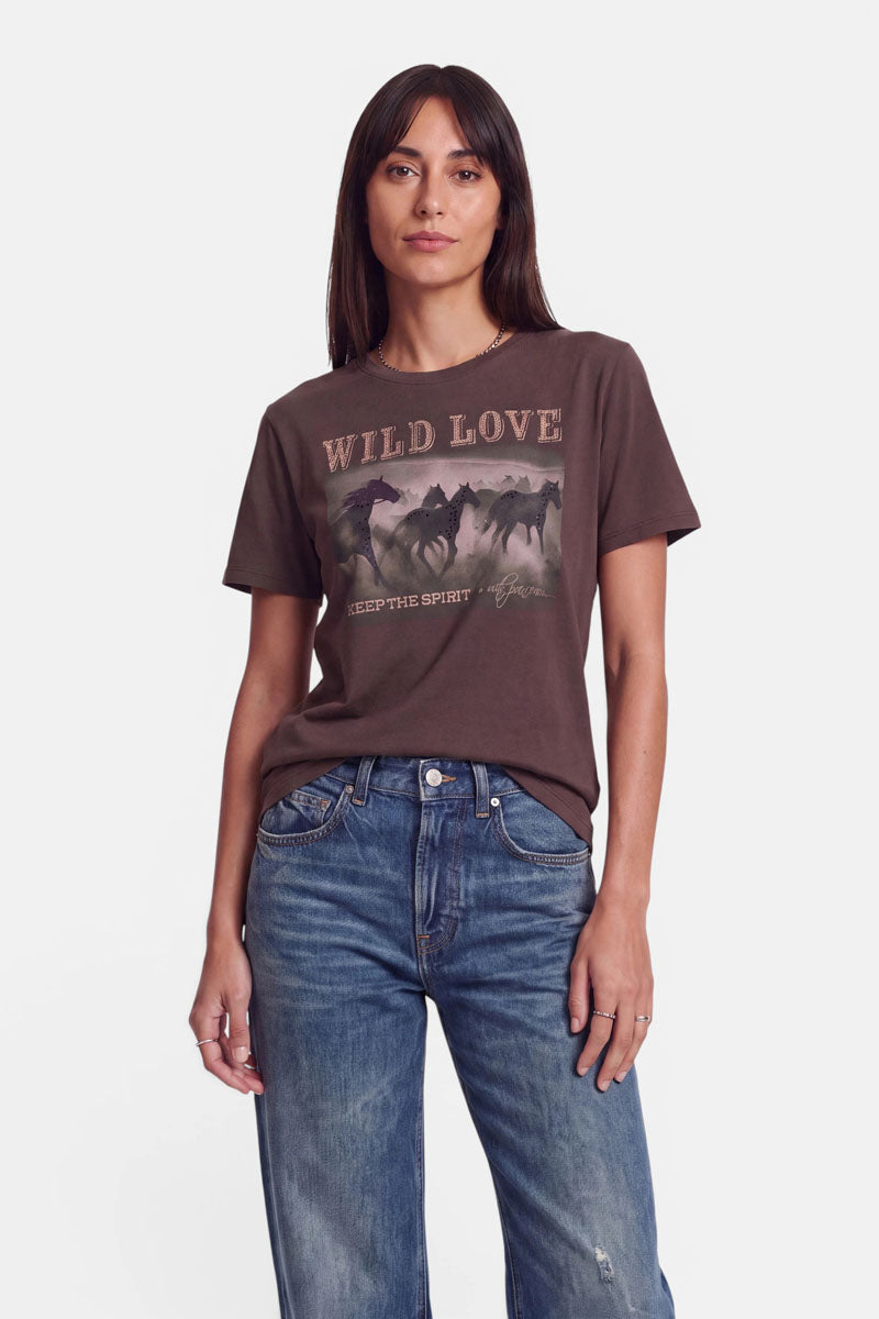 Eine Frau mit langen braunen Haaren trägt ein braunes T-Shirt mit der auffälligen Aufschrift „WILD LOVE“ und einem markanten Pferde-Print, dazu eine blaue Jeans. Sie steht mit dem Gesicht nach vorne vor einem schlichten weißen Hintergrund, mit neutralem Gesichtsausdruck und einer Hand in der Hosentasche.