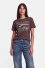 Eine Frau mit langen braunen Haaren trägt ein braunes T-Shirt mit der auffälligen Aufschrift „WILD LOVE“ und einem markanten Pferde-Print, dazu eine blaue Jeans. Sie steht mit dem Gesicht nach vorne vor einem schlichten weißen Hintergrund, mit neutralem Gesichtsausdruck und einer Hand in der Hosentasche.