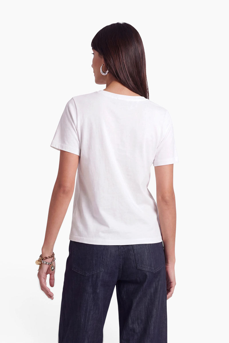Eine Frau mit glattem braunem Haar steht mit dem Rücken zur Kamera. Sie trägt Creolen, ein weißes Baumwoll-T-Shirt mit kurzen Ärmeln, eine dunkle Jeans mit hoher Taille und goldene Armbänder. Der Hintergrund ist schlicht weiß.