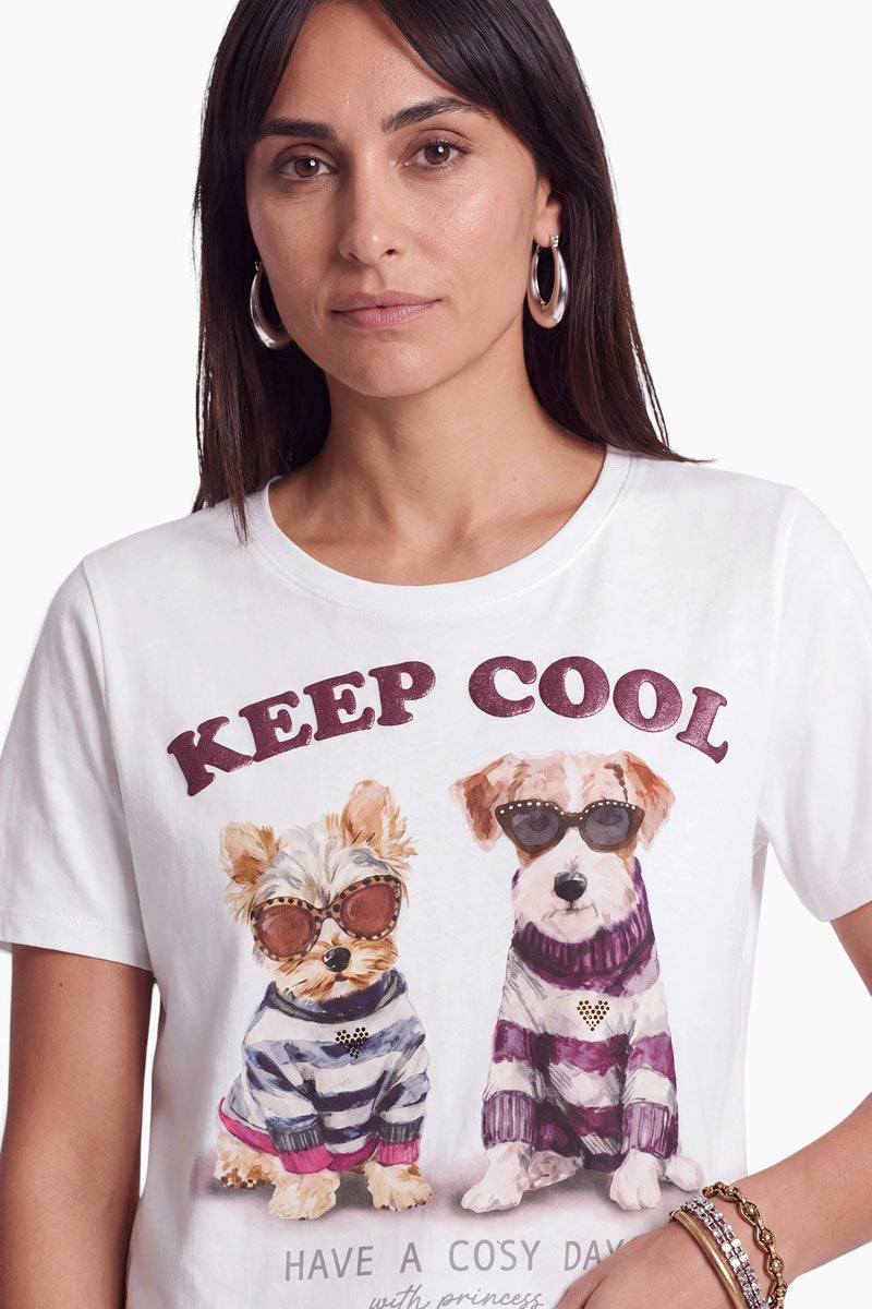 Eine Frau mit langen dunklen Haaren trägt ein weißes Baumwoll-T-Shirt mit einem lustigen Hundemotiv: zwei Hunde in Pullovern und Sonnenbrillen, darüber der Schriftzug „KEEP COOL“ und darunter ein teilweise sichtbarer Text. Sie trägt Creolen und Armbänder und blickt vor einem einfarbigen Hintergrund direkt in die Kamera.