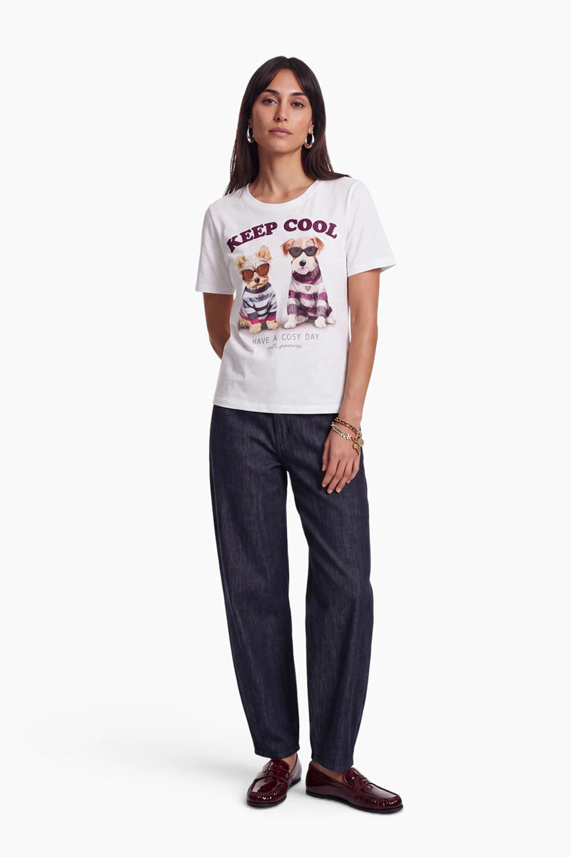 Eine Frau steht vor einem weißen Hintergrund und trägt ein weißes Baumwoll-T-Shirt mit einem niedlichen Hundemotiv, auf dem Comic-Hunde in Pullovern zu sehen sind, sowie den Schriftzug „KEEP COOL“. Dazu trägt sie eine weite dunkle Hose und burgunderrote Loafer. Sie hat langes dunkles Haar, Creolen und Strassarmbänder, die ihrem lässigen Look einen Hauch von Glanz verleihen.