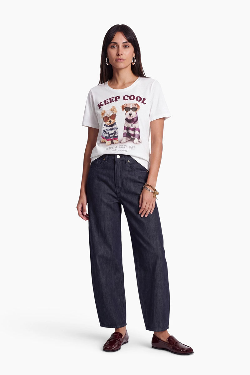 Eine Frau steht in einer entspannten Haltung da und trägt ein weißes Baumwoll-T-Shirt mit einem Hunde-Aufdruck – zwei gezeichnete Hunde –, eine lockere dunkle Jeans, braune Slipper, goldene Creolen und Armbänder. Sie hat glattes dunkles Haar und einen neutralen Gesichtsausdruck. Schlichter weißer Hintergrund.