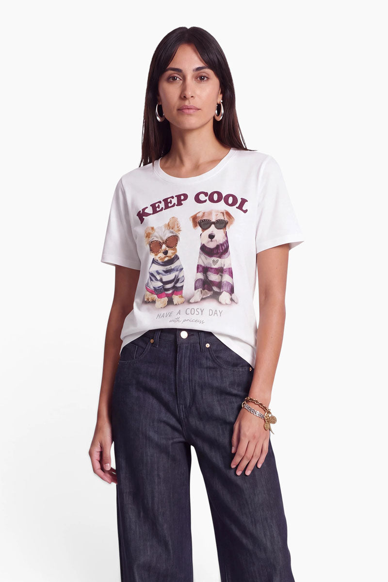 Eine Frau mit langen, dunklen Haaren trägt Creolen, ein Baumwoll-T-Shirt mit Hundemotiv, auf dem zwei Hunde in Pullovern und der Schriftzug „KEEP COOL“ zu sehen sind, dazu eine dunkle Jeans mit weitem Bein. Sie steht mit ausdruckslosem Gesicht vor einem schlichten weißen Hintergrund, wobei ihr dezenter Stil durch einen Hauch von Strass betont wird.
