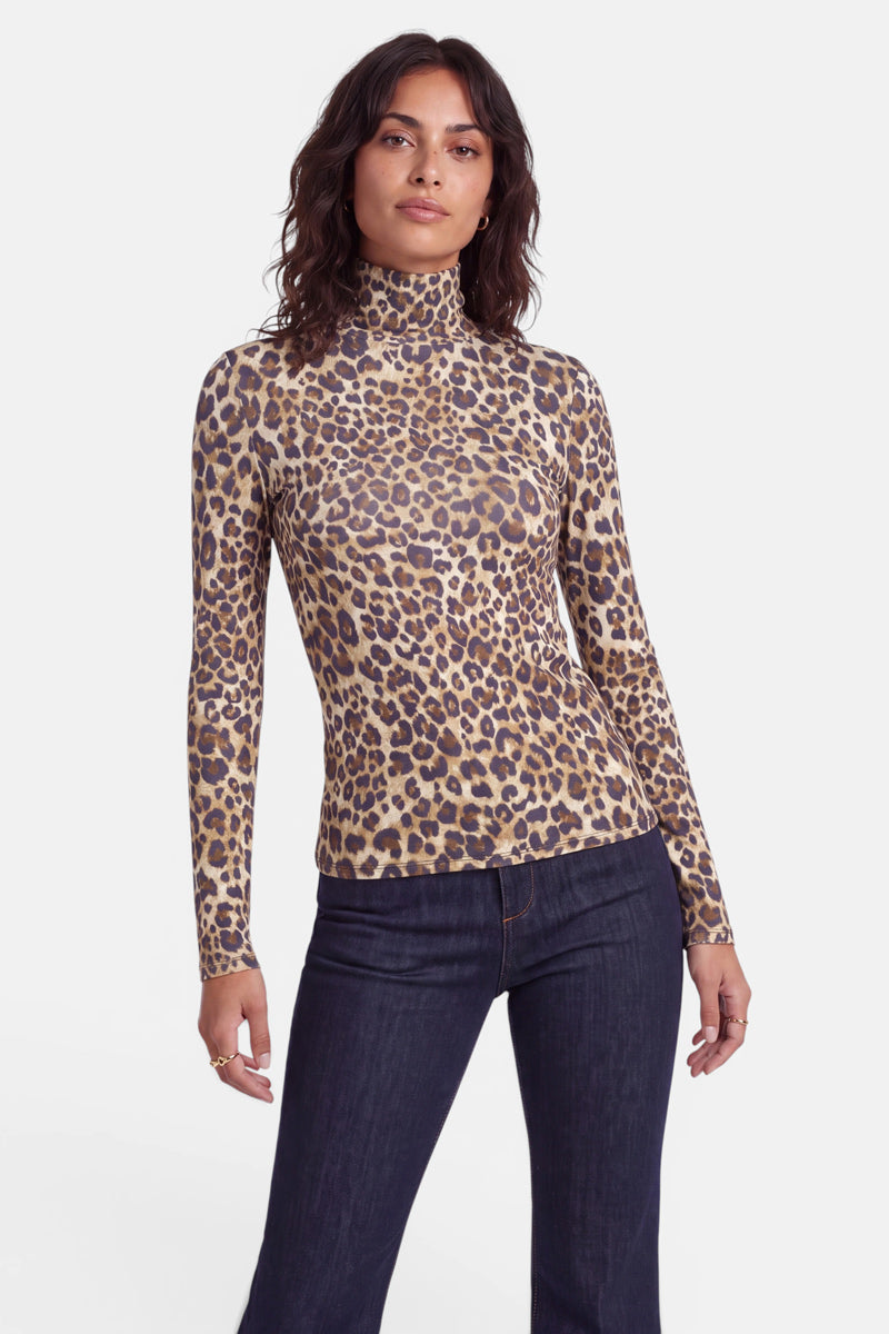 Eine Person mit welligem, dunklem Haar trägt ein eng anliegendes, langärmeliges Langarmshirt mit Leopardenmuster und Stehkragen sowie eine dunkle Jeans. Sie steht vor einem schlichten weißen Hintergrund, blickt mit neutralem Gesichtsausdruck und entspannter Körperhaltung nach vorne.