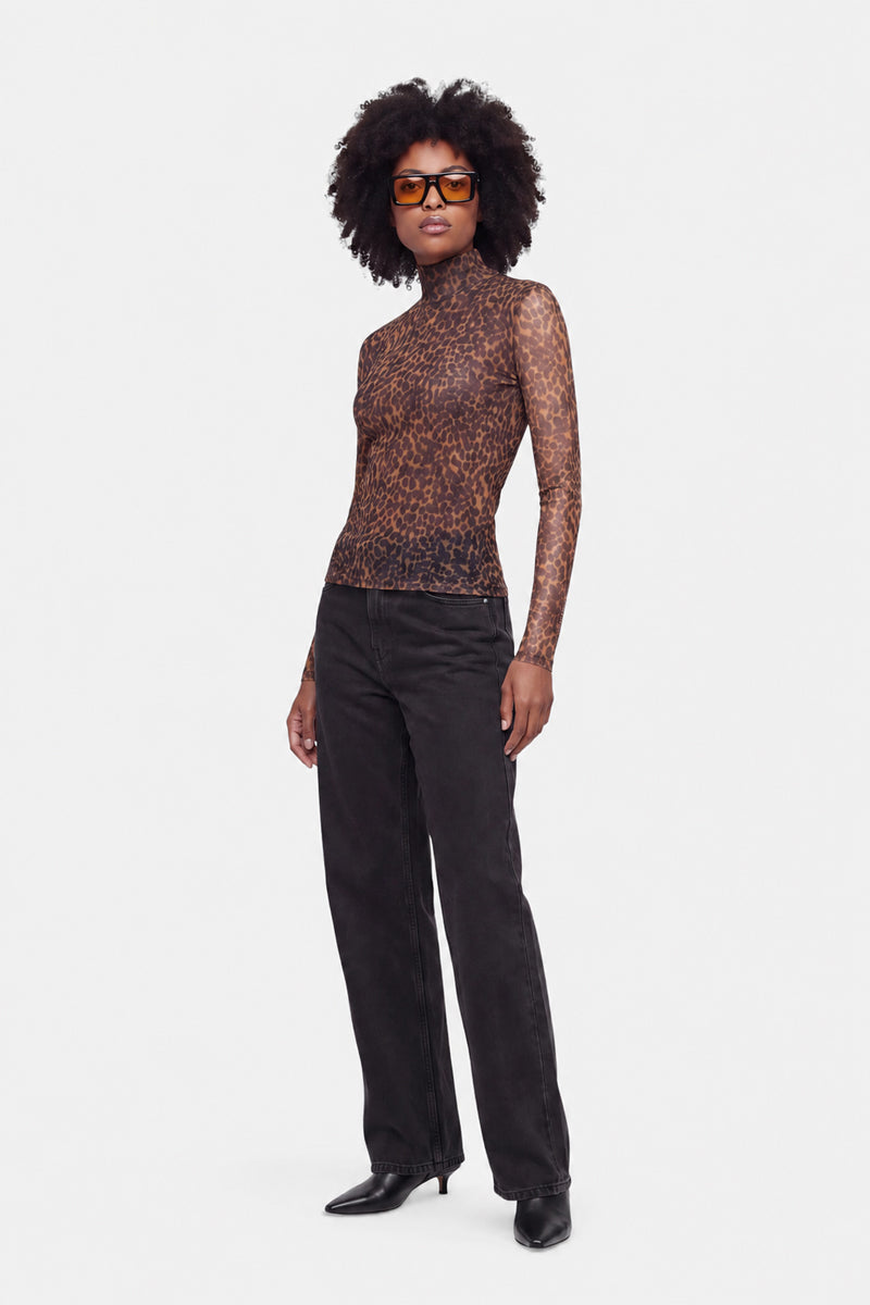 Eine Frau mit Afro-Frisur trägt eine braune Sonnenbrille, ein halbtransparentes Langarmshirt aus Netzstoff mit Leo-Print, eine weite schwarze Hose und schwarze spitze Schuhe. Sie steht vor einem schlichten weißen Hintergrund.