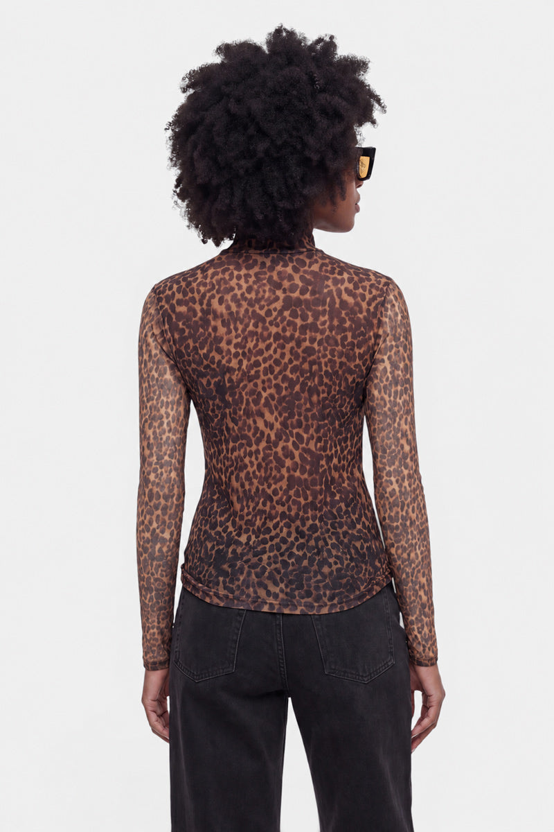 Eine Person mit lockigem Haar, die eine Sonnenbrille, ein transparentes Langarmshirt aus Netzstoff mit Leo-Print und schwarze Jeans trägt, steht mit dem Rücken zur Kamera vor einem schlichten, hellen Hintergrund.