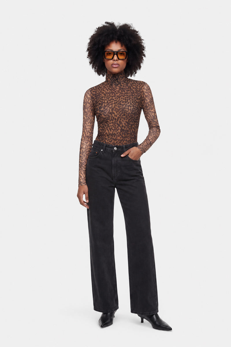 Eine Person steht vor einem weißen Hintergrund und trägt ein durchsichtiges Langarmshirt aus Mesh mit Löwenprint, eine schwarze Jeans mit hoher Taille, schwarze spitze Stiefel und eine große, eckige Sonnenbrille. Sie hat eine selbstbewusste Haltung und trägt eine natürliche Afro-Frisur.