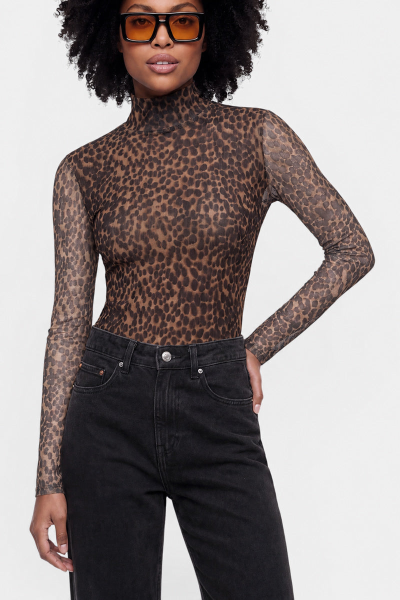 Eine Person mit natürlicher Afro-Frisur trägt eine große braune Sonnenbrille, ein eng anliegendes, langärmeliges Mesh-Turtleneck-Shirt mit Leo-Print (Langarmshirt) und eine schwarze Jeans mit hoher Taille und steht vor einem hellgrauen Hintergrund, wobei sie eine Hand in die Hüfte gestützt hat.
