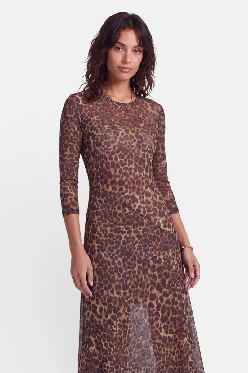 Eine Frau mit welligem, dunklem Haar trägt ein figurbetontes, langärmeliges, braunes Midikleid mit Leopardenmuster. Das halbtransparente Netzkleid hat einen Rundhalsausschnitt und eine A-Linien-Silhouette. Sie steht vor einem schlichten weißen Hintergrund, blickt nach vorne und hat einen neutralen Gesichtsausdruck.