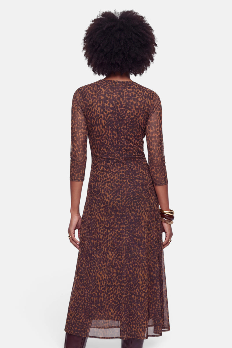 Eine Frau steht mit dem Rücken zur Kamera und trägt ein transparentes, figurbetontes Meshkleid mit Dreiviertelärmeln und knöchellanger Midilänge, das mit einem auffälligen braun-schwarzen Leo-Print verziert ist. Ihr Haar ist naturbelassen und voluminös, und am rechten Handgelenk trägt sie mehrere goldene Armreifen. Der Hintergrund ist schlicht weiß.