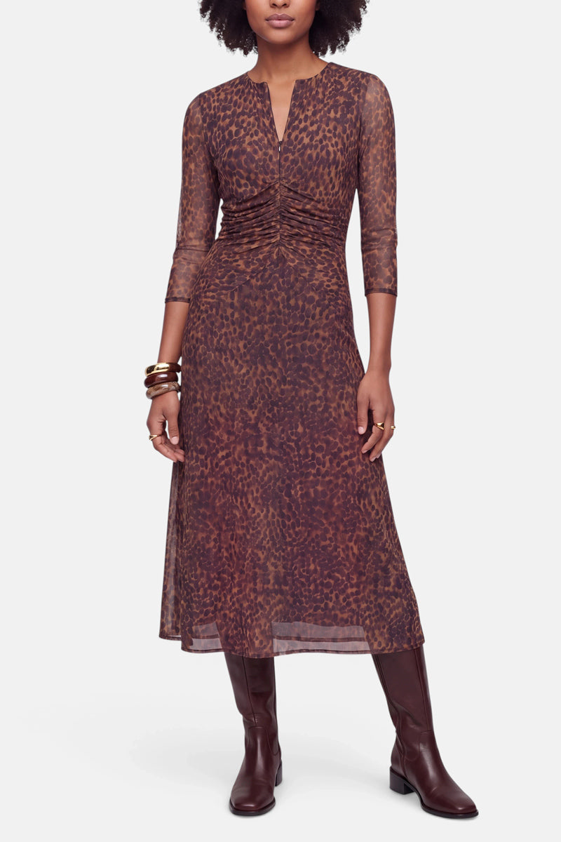 Eine Frau trägt ein braunes, langärmeliges Meshkleid mit einem abstrakten Leo-Print-Muster in Midilänge. Das Kleid ist an der Taille gerafft und hat einen kleinen Ausschnitt mit Schlitz vorne. Sie trägt dazu mehrere Armreifen und kniehohe braune Lederstiefel.