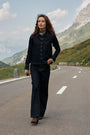 Eine Frau mit welligem braunem Haar geht selbstbewusst eine Bergstraße entlang. Sie trägt eine dunkle Jeansjacke und dazu passende Jeanshosen in dunkler Waschung mit Schlag, eine Perlenkette sowie Stiefel mit Absatz. Unter einem teilweise bewölkten Himmel sind grüne Hügel und schneebedeckte Gipfel zu sehen.