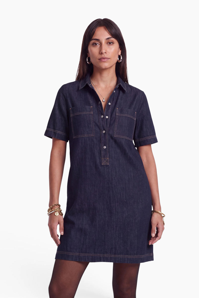 Eine Frau steht mit dem Blick nach vorne und trägt ein kurzärmeliges, dunkles Jeanskleid aus Denim mit Kragen, Druckknöpfen und zwei Brusttaschen. Das Kleid aus Baumwolle in einer dunklen Waschung passt perfekt zu ihrem glatten braunen Haar, den Creolen, Armbändern und Halsketten. Der Hintergrund ist schlicht weiß.