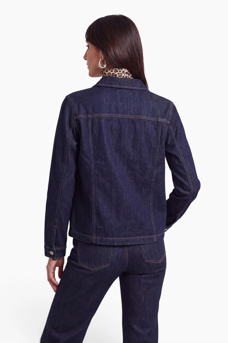 Eine Frau mit glattem, dunklem Haar steht mit dem Rücken zur Kamera und trägt eine dunkle Jeansjacke in dunkler Waschung sowie dazu passende Jeans. Ein Arm ist angewinkelt, und unter ihrem Jeanshemd aus Baumwolle lugt ein Kragen mit Leopardenmuster hervor. Der Hintergrund ist schlicht weiß.