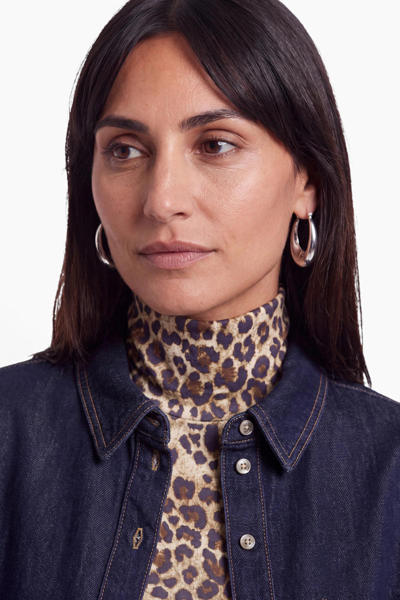 Eine Frau mit glattem, dunklem Haar trägt silberne Creolen, einen Rollkragenpullover mit Leopardenmuster und ein dunkles Jeanshemd aus Baumwolle in dunkler Waschung. Vor einem schlichten weißen Hintergrund blickt sie mit neutralem Gesichtsausdruck leicht zur Seite.