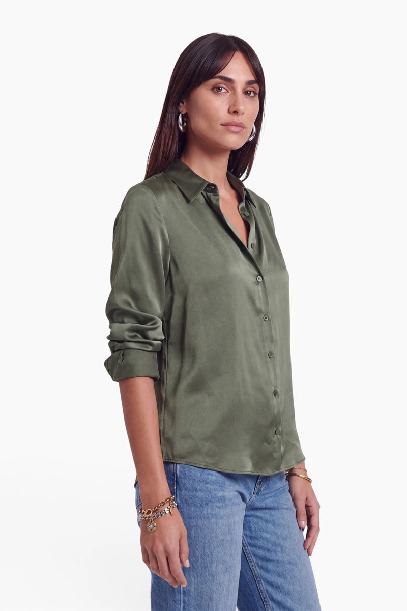 Eine Frau trägt eine olivgrüne, langärmelige Stretch-Hemdbluse, die sie in ihre blaue Jeans gesteckt hat. Sie steht seitlich und hat die Ärmel bis zu den Ellbogen hochgekrempelt.