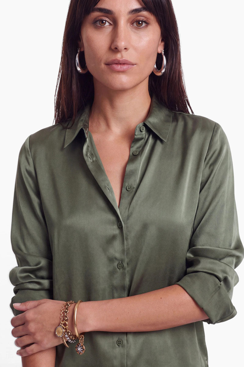 Eine Frau trägt eine olivgrüne Stretch-Hemdbluse und silberne Creolen. Sie posiert mit verschränkten Armen und zeigt dabei ein mehrreihiges Goldarmband an ihrem Handgelenk.